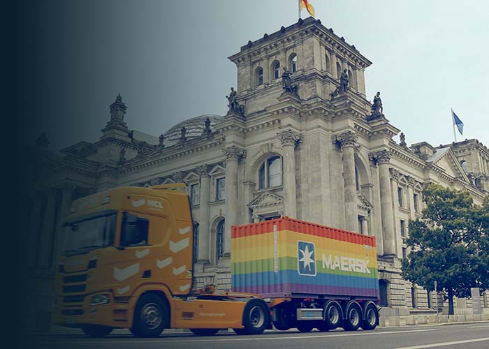 A truck carrying the Maersk rainbow container on road with a building in the background.