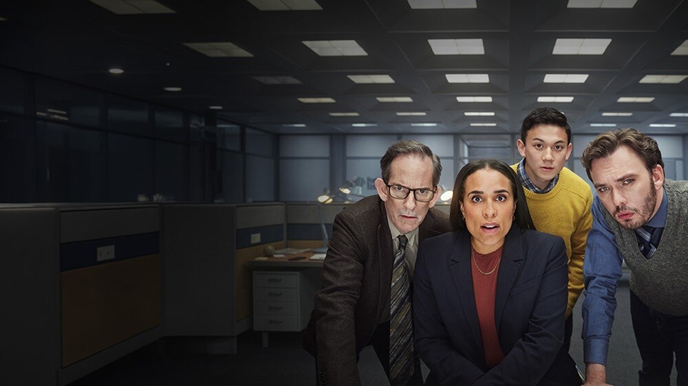Four office employees intensely looking at a computer screen in a dimly lit modern office space