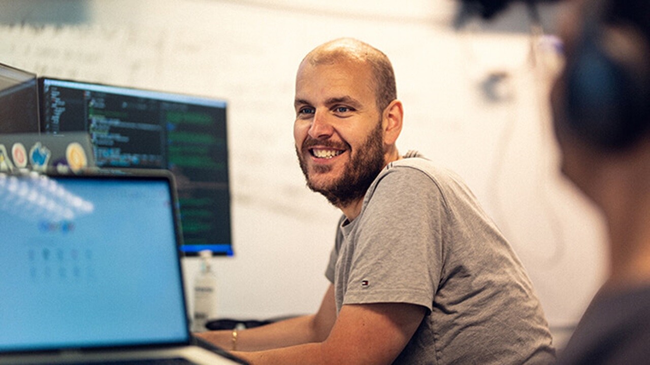 An architect at his workstation smiling and talking to one his fellow architects.