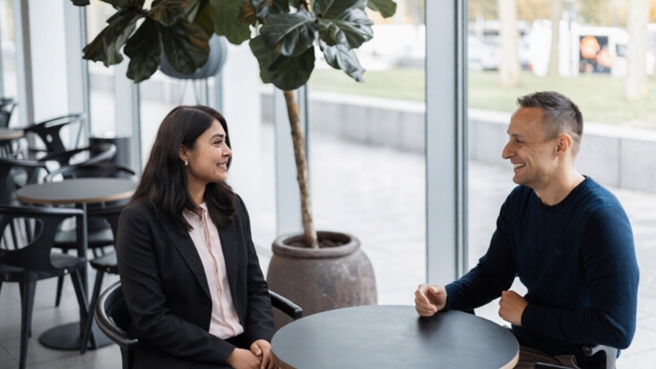 Two colleagues seated at a small table in a bright office lounge, engaged in conversation.