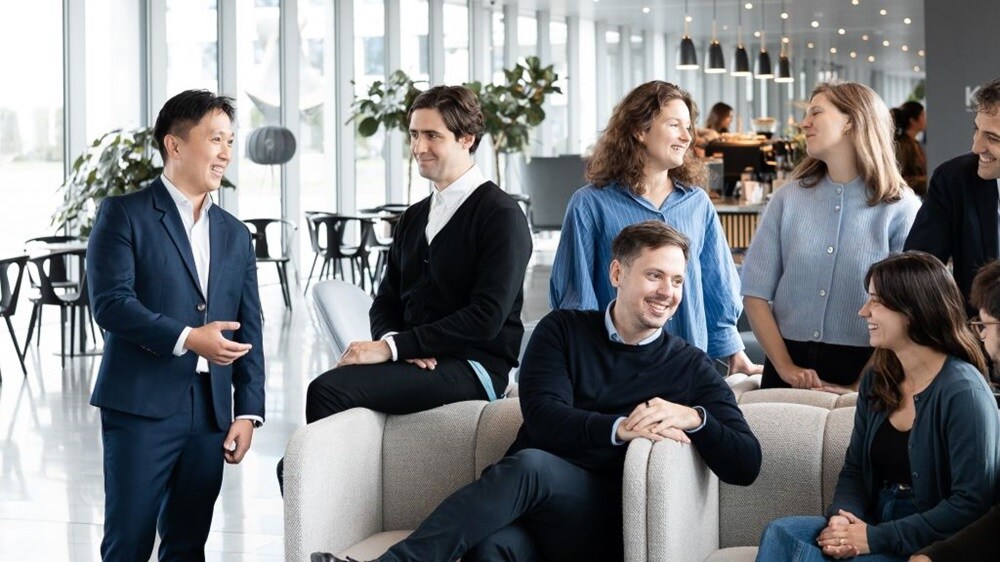 Group of colleagues gathered in a modern office lounge area, engaged in conversation with open seating, plants, and workspaces in the background.