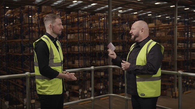 Two men in safety vests converse in a warehouse, surrounded by shelves and boxes.