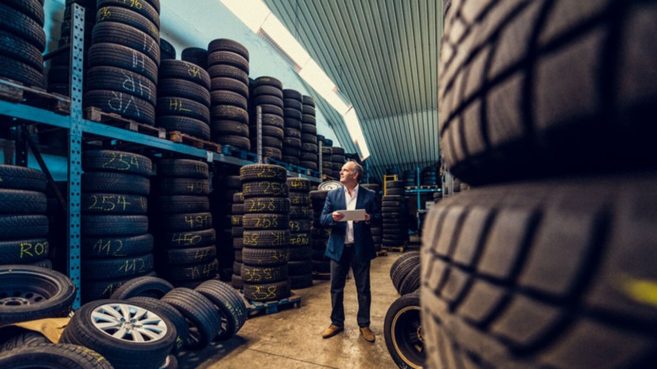 A man wearing a suit stands in a storage facility surrounded by towers of car tyres