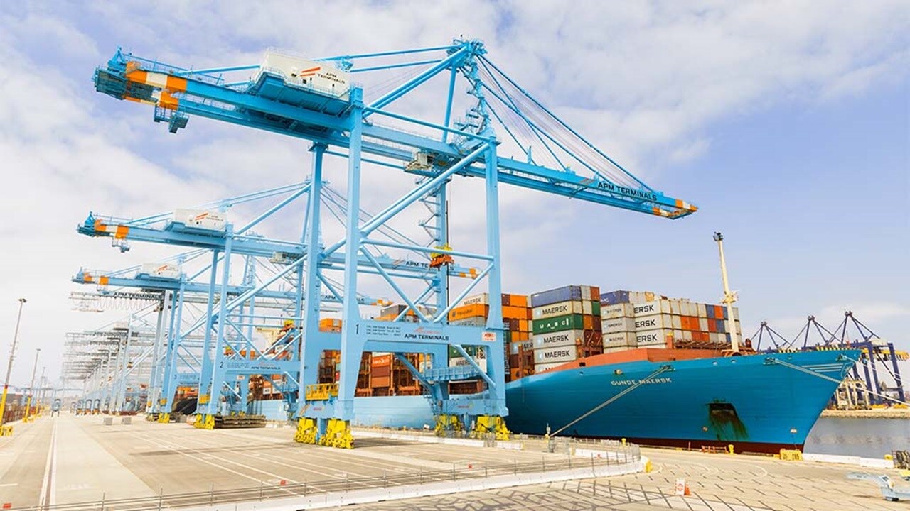 A large container ship is docked at a busy port, with cranes and cargo containers visible in the background.