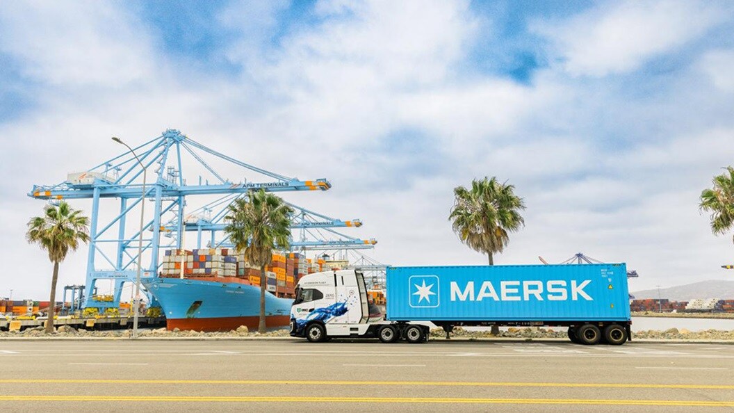 A Maersk container truck parked at APM Terminals with a large container ship and port cranes in the background.