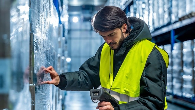 A man wearing a safety vest scans items with a handheld device in a warehouse setting.