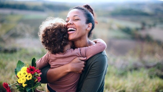 A woman and her child embrace warmly in a sunlit field, surrounded by tall grass and wildflowers.