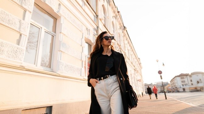 A woman in a black coat and white pants walks down a city street.