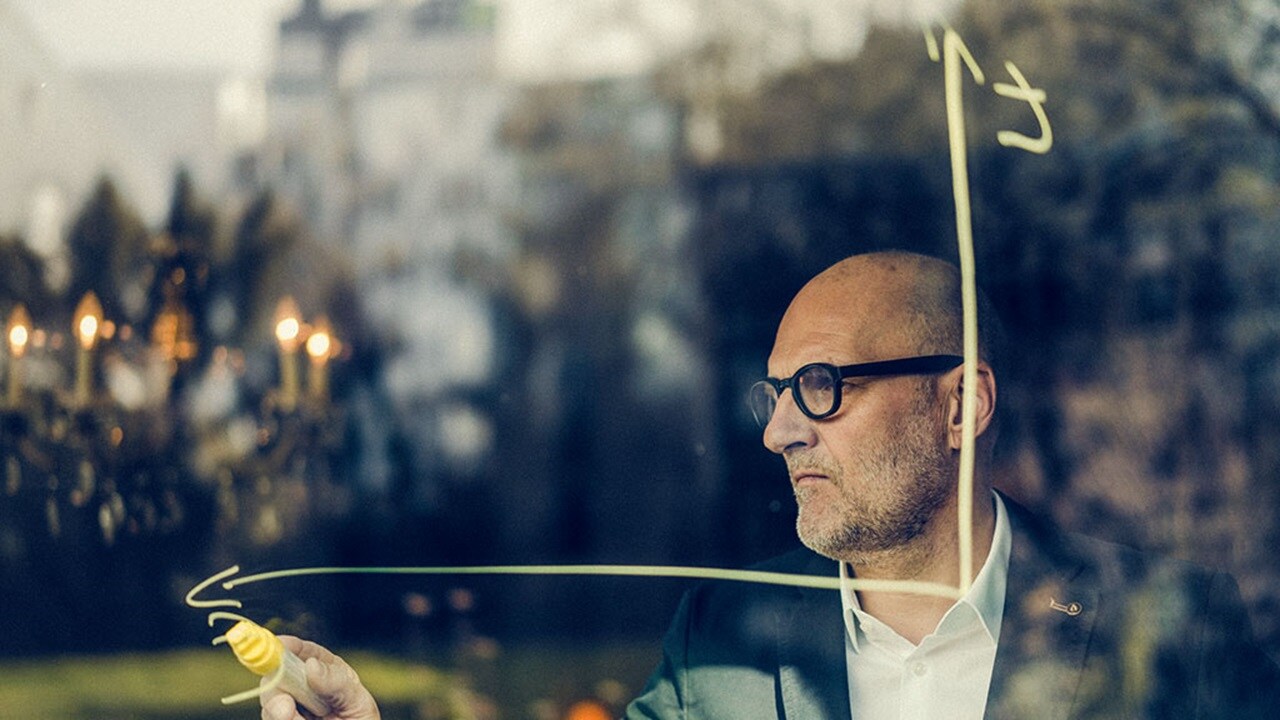 A man in a suit and glasses sketches on a glass window, focused on his drawing.