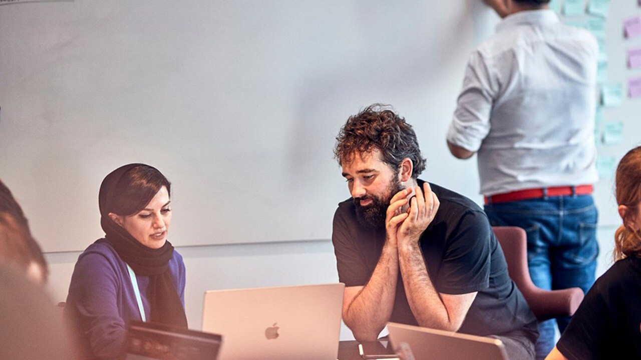 Two people sitting at a desk in a workshop, looking at a laptop