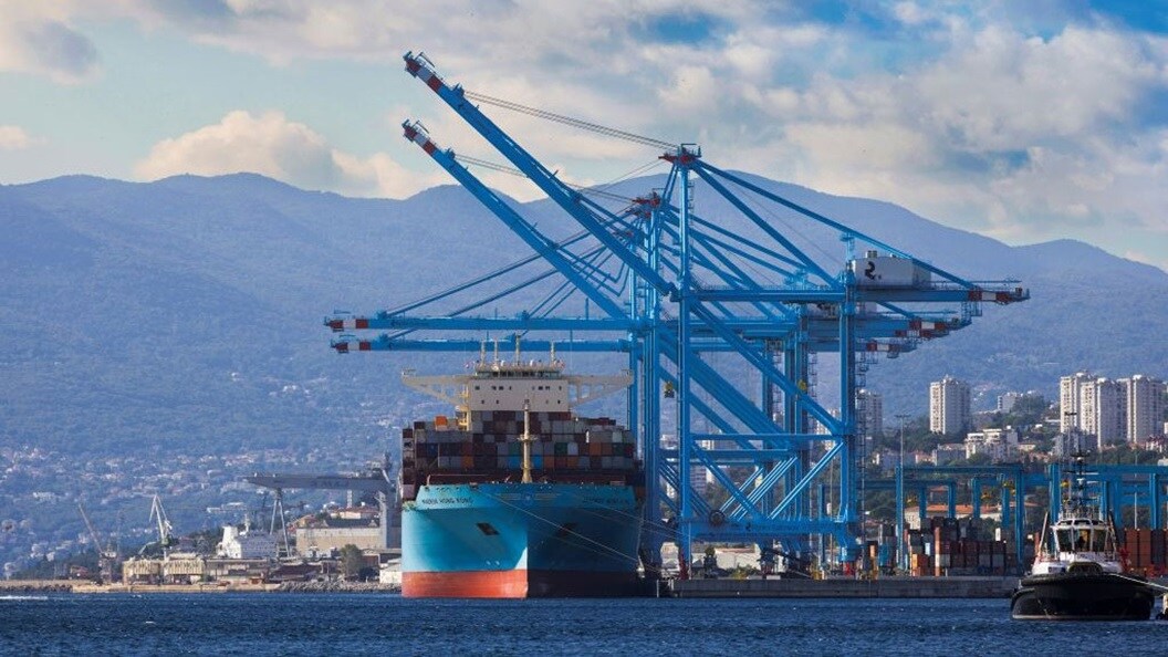 Large cargo ship docked at a port with blue container cranes, mountains in the background, and a partly cloudy sky.