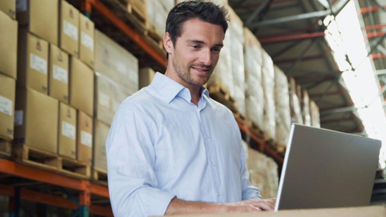 A smiling man working on laptop in a cargo warehouse.