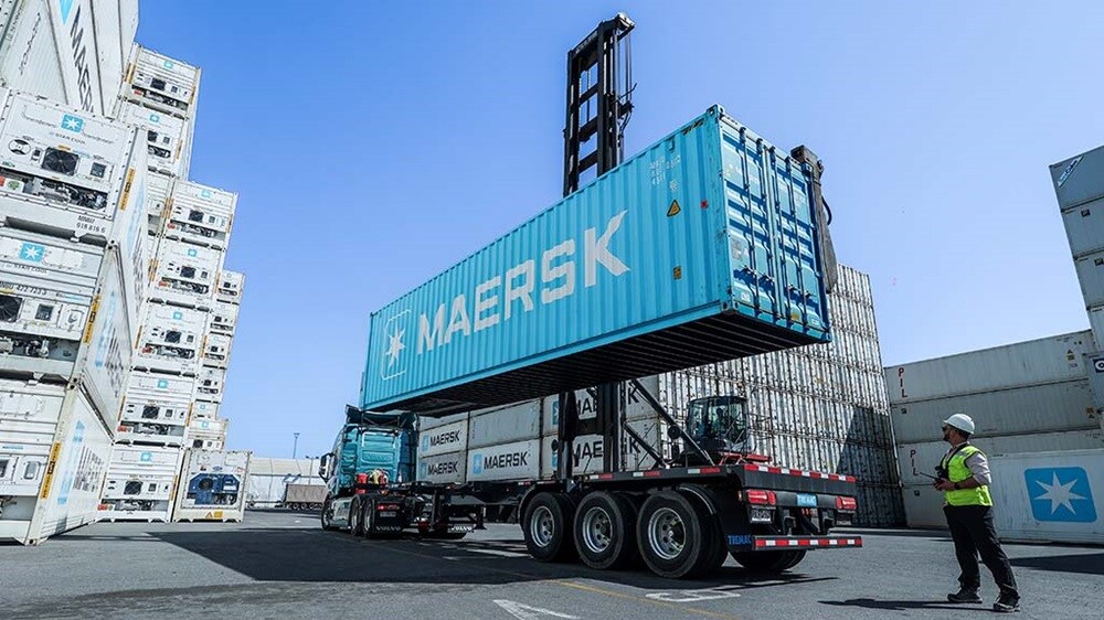 Maersk shipping container being lifted onto a truck by a reach stacker in a container yard, with stacked containers and a port worker nearby.
