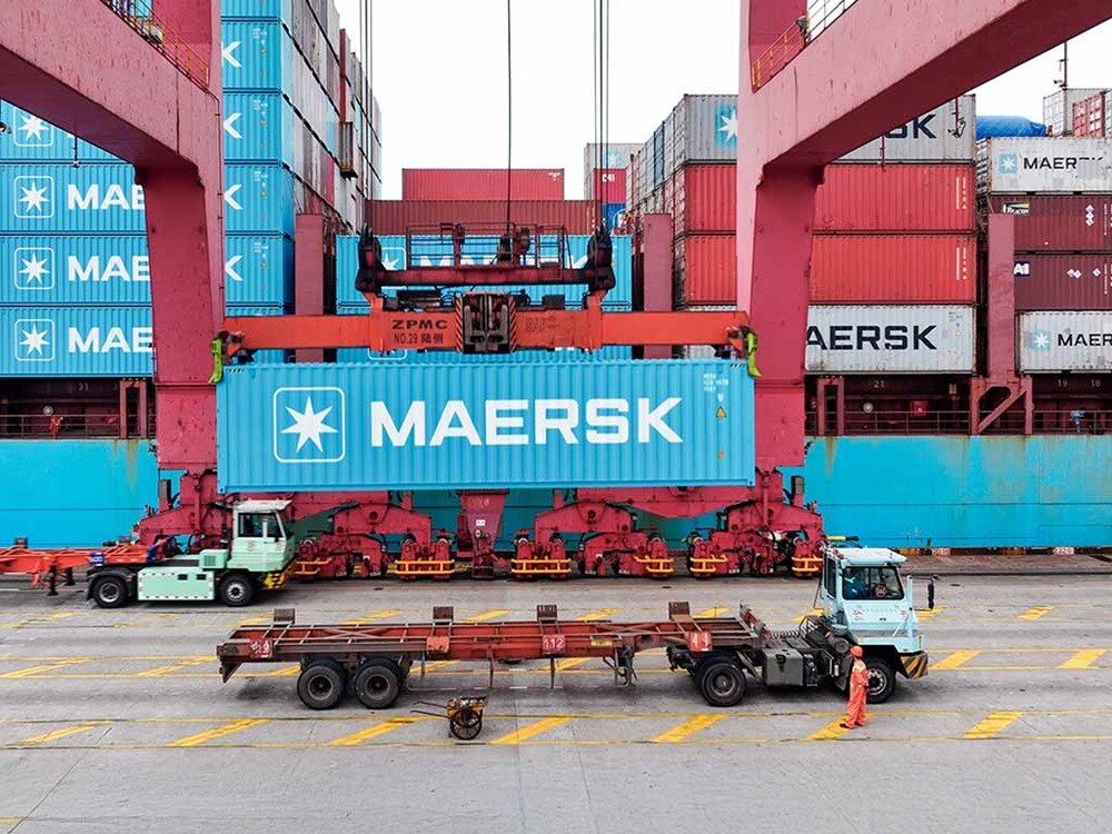 Yoğun bir liman terminalinde altında kamyonların bulunduğu bir gezer köprülü vinç ile kaldırılan Maersk nakliye konteyneri