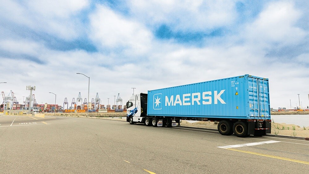 A blue Maersk truck is parked on a road near an industrial port, with shipping containers and cranes visible in the background under a cloudy sky.